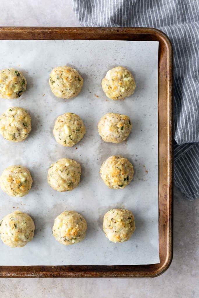 Twelve uncooked, evenly spaced dough balls with herbs on a parchment-lined baking sheet, next to a blue and white striped cloth.