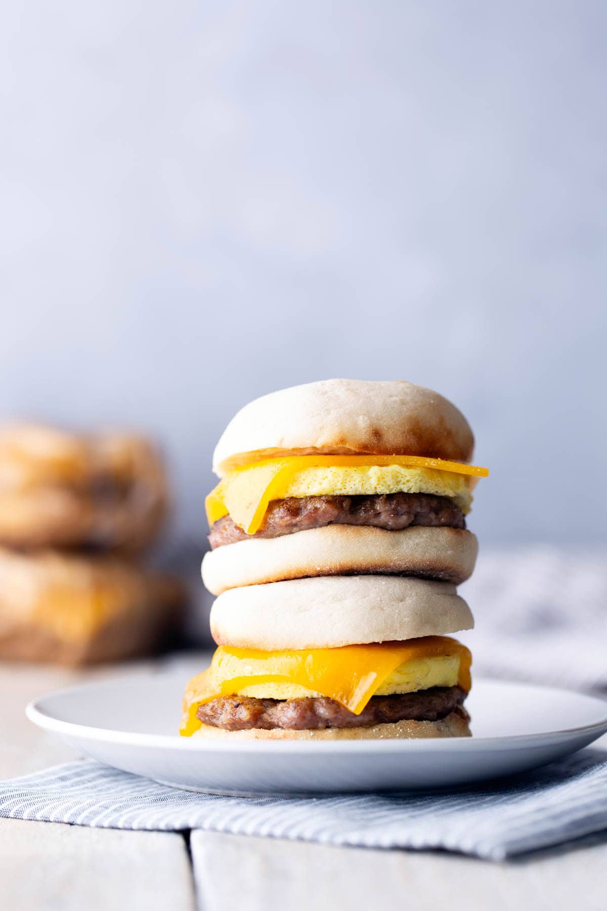 Two breakfast sandwiches with egg, sausage, and cheddar cheese are stacked on a white plate, with more sandwiches blurred in the background.
