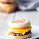 Breakfast sandwich with sausage, egg, and cheddar cheese on an English muffin, served on a white plate with a striped cloth beneath.