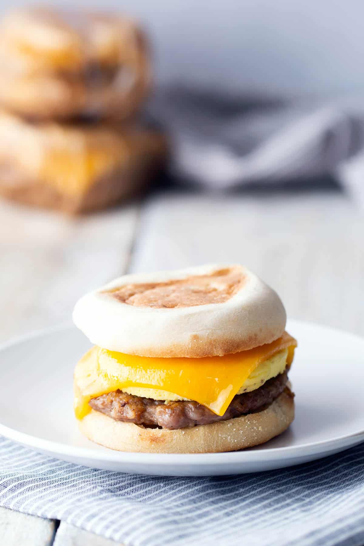 Breakfast sandwich with sausage, egg, and cheddar cheese on an English muffin, served on a white plate with a striped cloth beneath.