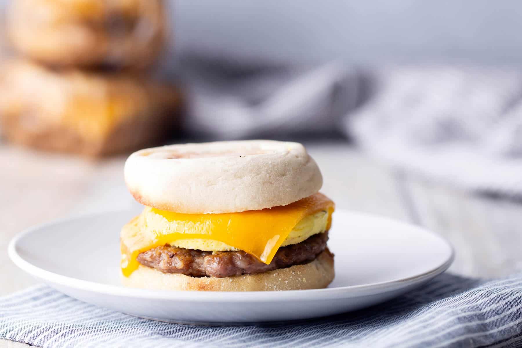 A breakfast sandwich with egg, cheddar cheese, and sausage inside an English muffin sits on a white plate.