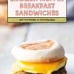 A breakfast sandwich with egg, cheddar cheese, and sausage on an English muffin sits on a white plate. Text above reads: "English Muffin Breakfast Sandwiches. Get the recipe at stetted.com.