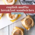 English muffin breakfast sandwiches with cheese, some unwrapped on a white surface and some wrapped in plastic and string on a light wooden table with a striped cloth nearby.