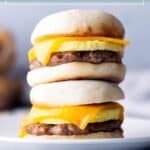 Two breakfast sandwiches with eggs, sausage patties, and cheddar cheese on English muffins, stacked on a white plate. Text at top reads "make-ahead breakfast sandwiches.