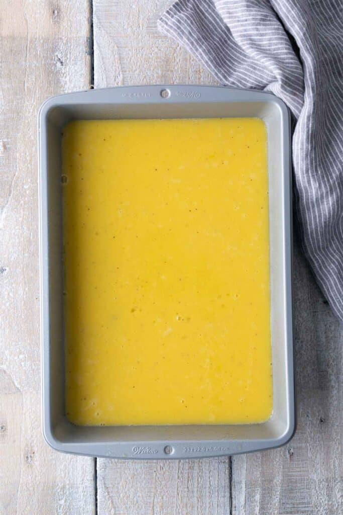 A rectangular metal baking pan filled with uncooked yellow liquid batter, placed on a light wooden surface next to a gray striped cloth.