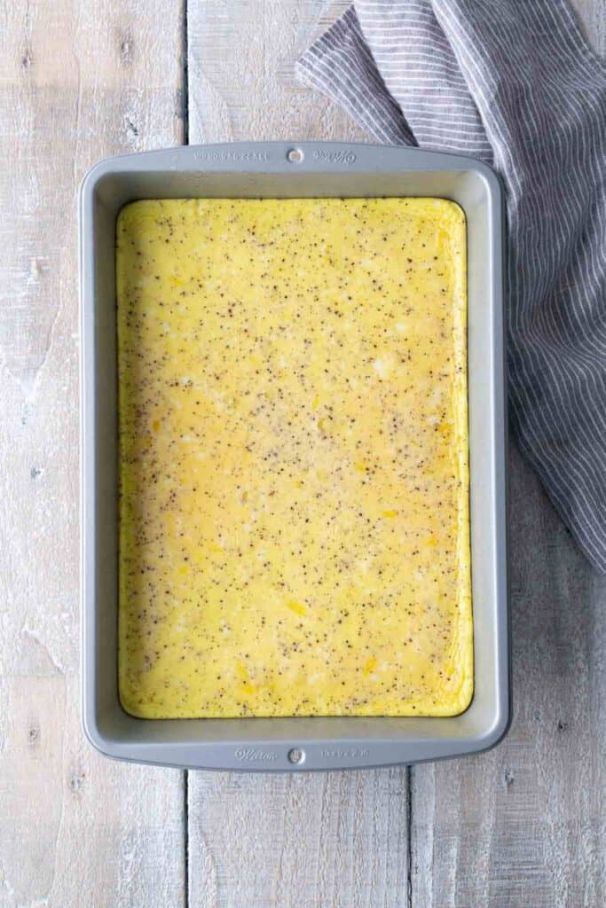 A rectangular metal baking pan filled with an uncooked egg mixture sits on a light wooden surface next to a gray and white striped cloth.
