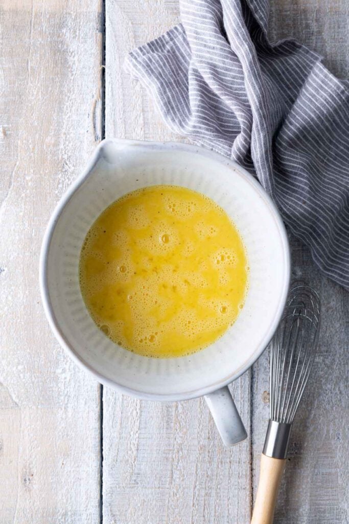 A white mixing bowl with whisked eggs next to a metal whisk and a striped cloth on a light wooden surface.