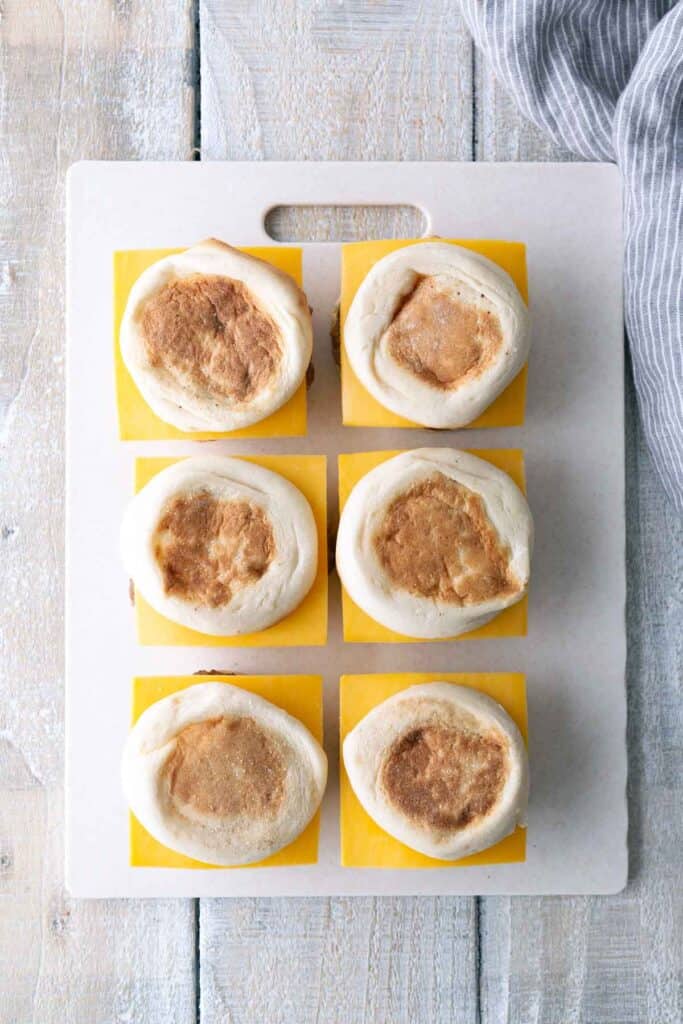 Six uncooked English muffins on square pieces of parchment paper are arranged in two rows of three on a white cutting board beside a striped cloth.