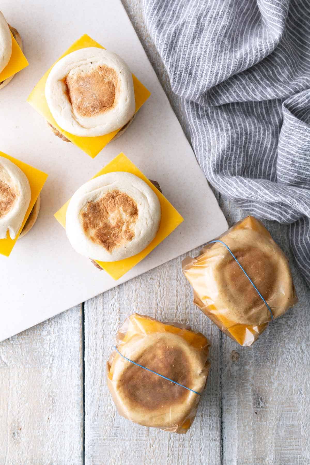 Six breakfast sandwiches with English muffins and cheese slices, some wrapped in wax paper and tied with string, are arranged on a white wooden surface next to a striped cloth.