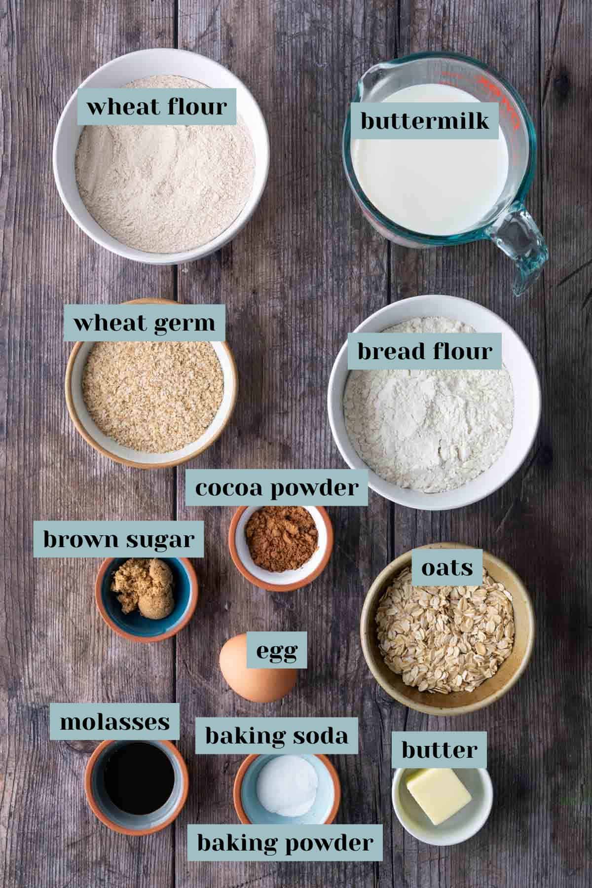 Top-down view of labeled baking ingredients in bowls and cups on a wooden surface, including flours, buttermilk, egg, oats, brown sugar, molasses, butter, and cocoa powder.
