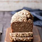 A loaf of oat-topped bread with two slices cut sits on a wooden cutting board, with a knife and a blue cloth in the background.