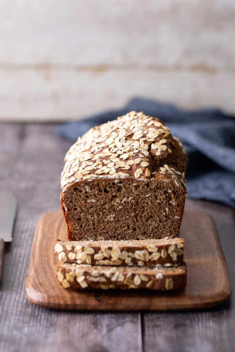 A loaf of oat-topped bread with two slices cut sits on a wooden cutting board, with a knife and a blue cloth in the background.