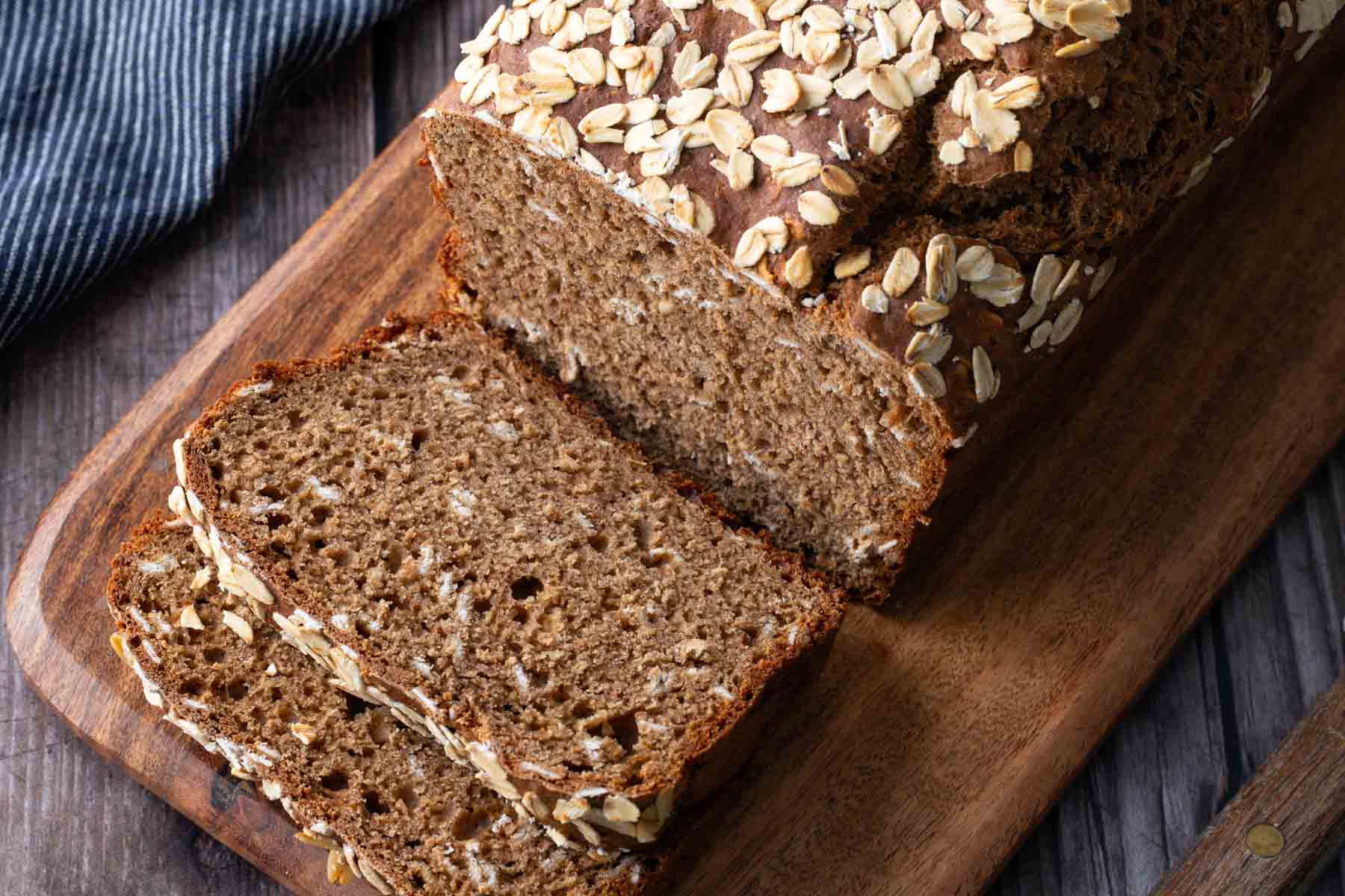 A loaf of brown oat bread partially sliced on a wooden cutting board, with oat flakes sprinkled on top.