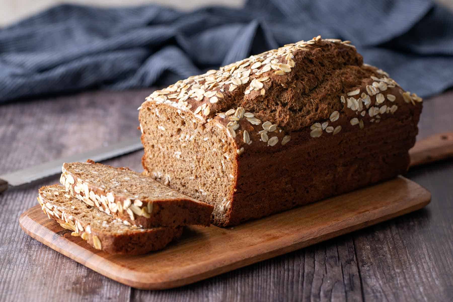 A loaf of whole grain bread topped with oats sits on a wooden cutting board with two slices cut, next to a bread knife on a rustic table.