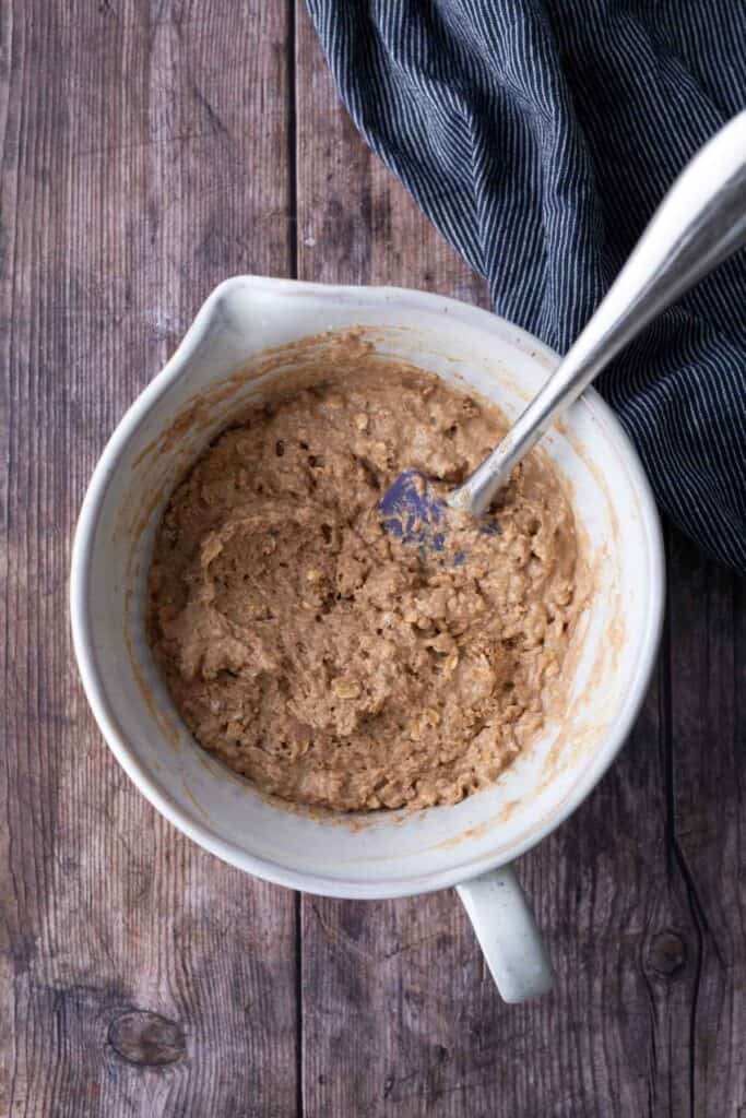 A mixing bowl filled with thick, brown batter and a spatula, placed on a wooden surface next to a dark striped cloth.