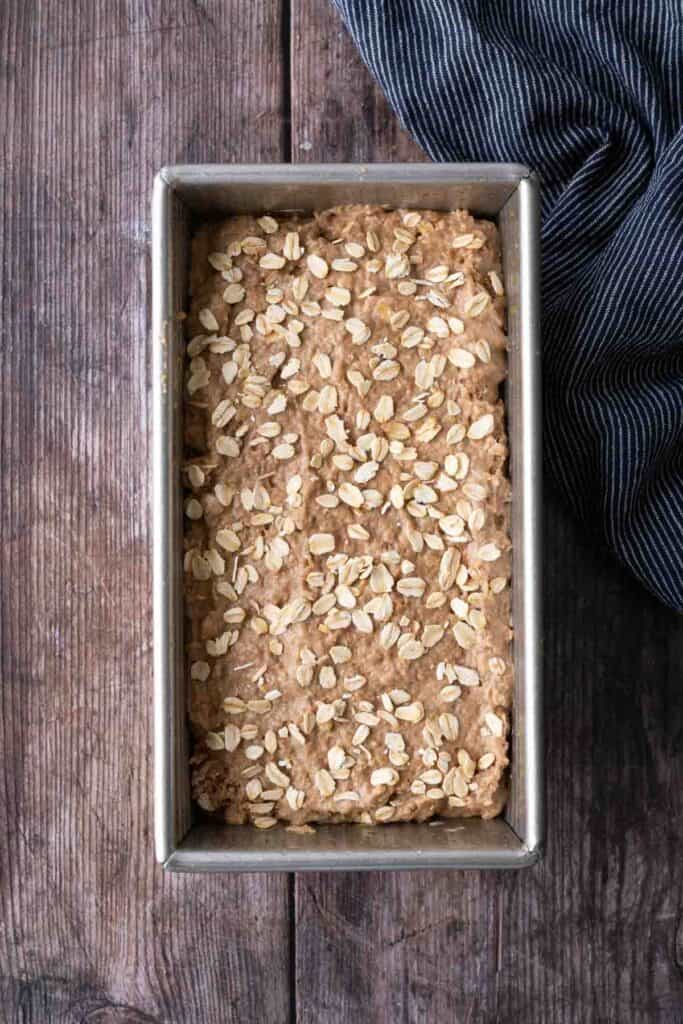 A metal loaf pan filled with unbaked oat-topped bread dough sits on a wooden surface next to a dark striped cloth.