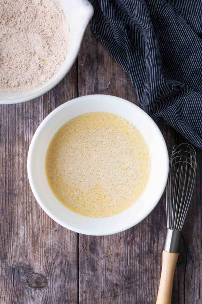 A white bowl with beaten eggs and milk sits on a wooden table next to a whisk, a dark cloth, and a bowl of flour mixture.