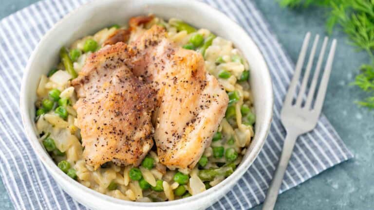 A bowl of creamy orzo pasta mixed with green peas, topped with two seasoned, cooked chicken thighs; a fork rests nearby on a striped napkin.