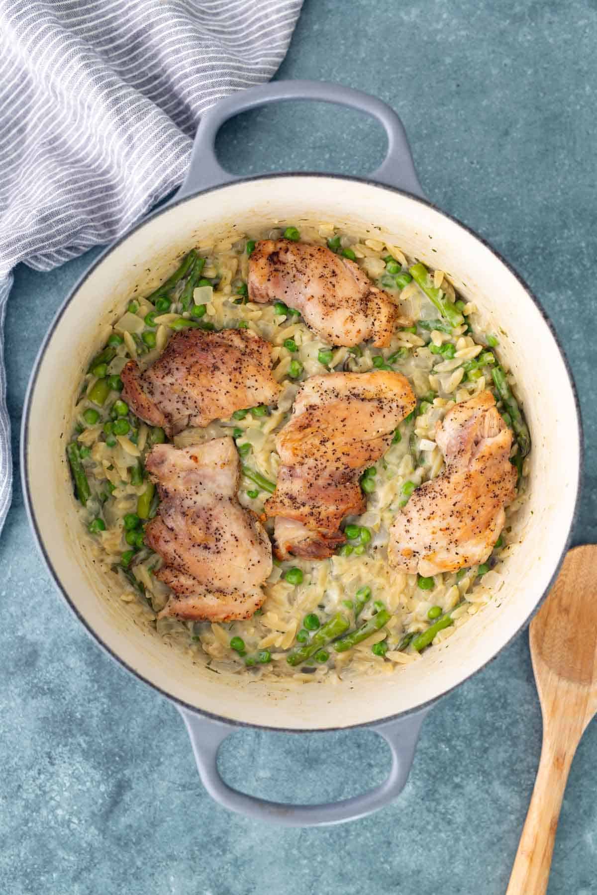 A Dutch oven filled with cooked chicken thighs on a bed of creamy rice, peas, and green beans, with a wooden spoon and striped towel nearby.