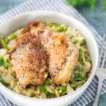 A bowl of creamy risotto with green peas, topped with two pieces of seasoned, cooked chicken, placed on a striped cloth with a fork beside it.