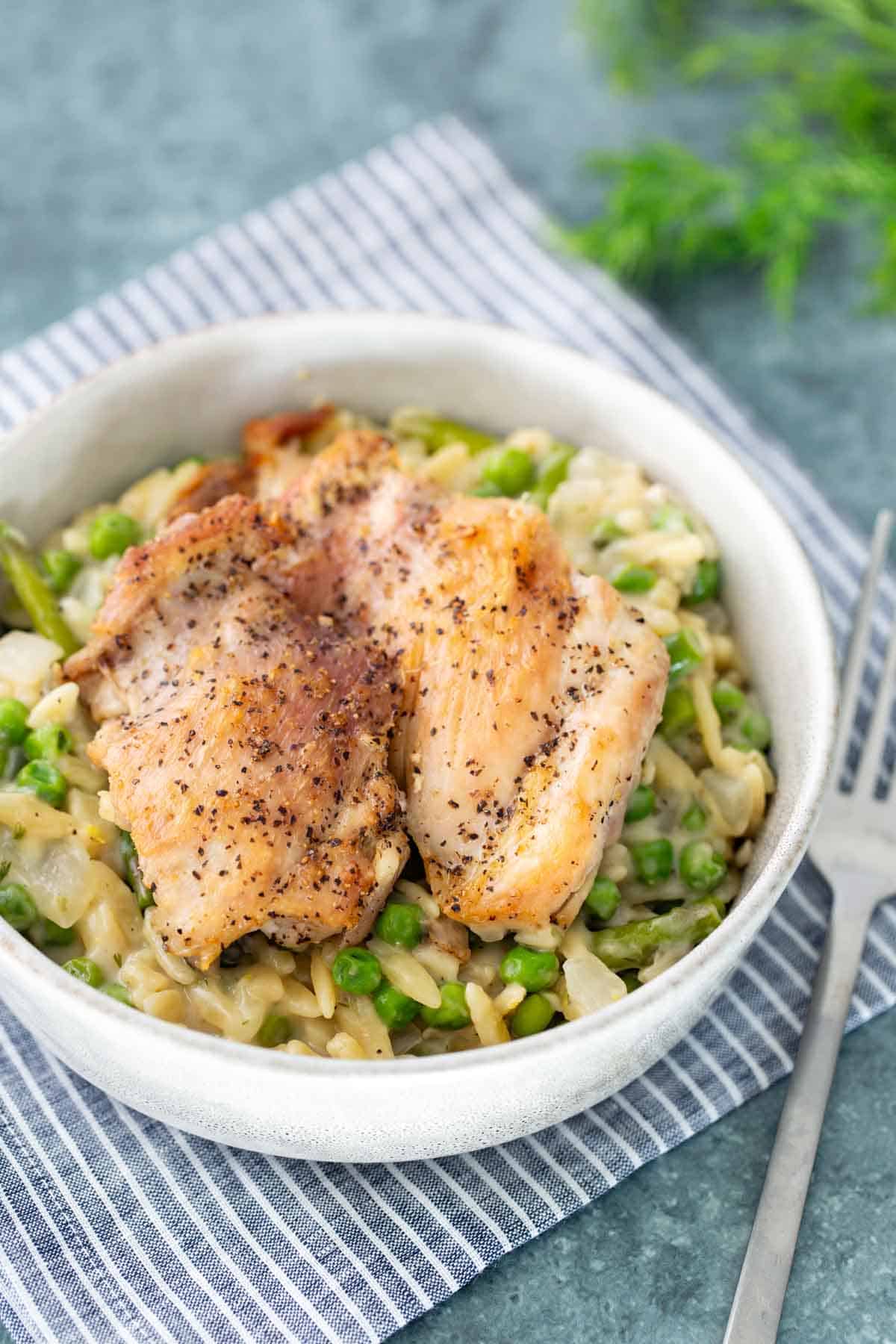 A bowl of creamy risotto with green peas, topped with two pieces of seasoned, cooked chicken, placed on a striped cloth with a fork beside it.