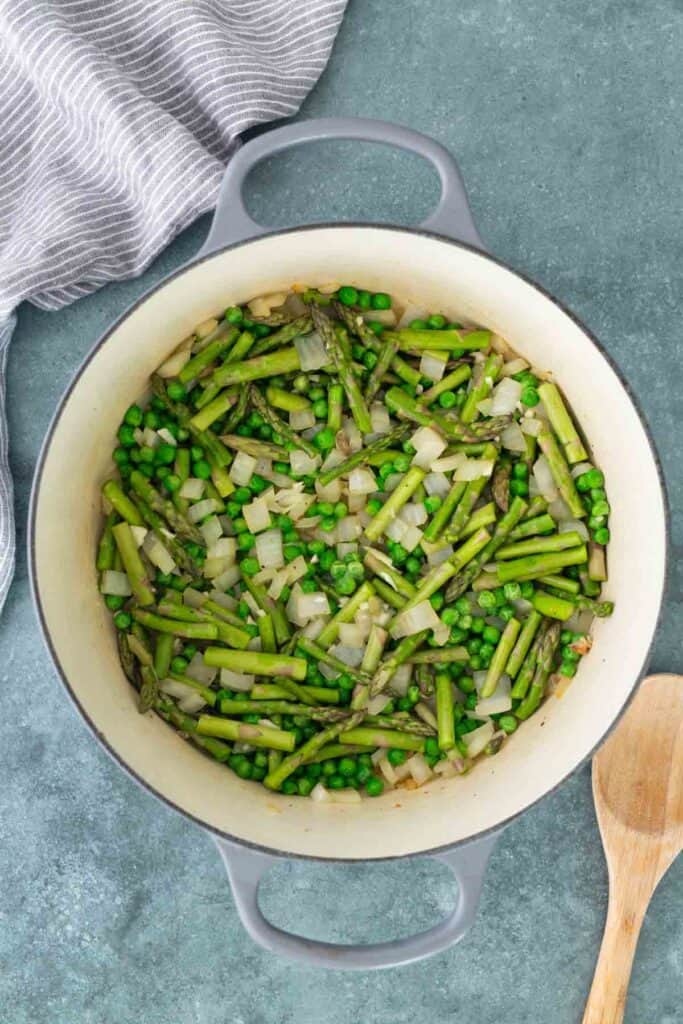 Chopped asparagus, peas, and onions cooking in a large gray pot on a green countertop, with a wooden spoon and striped towel nearby.