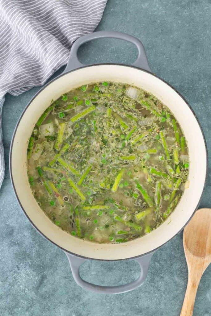 A pot of vegetable soup with chopped asparagus, onion, and herbs, sitting on a green countertop beside a striped towel and a wooden spoon.