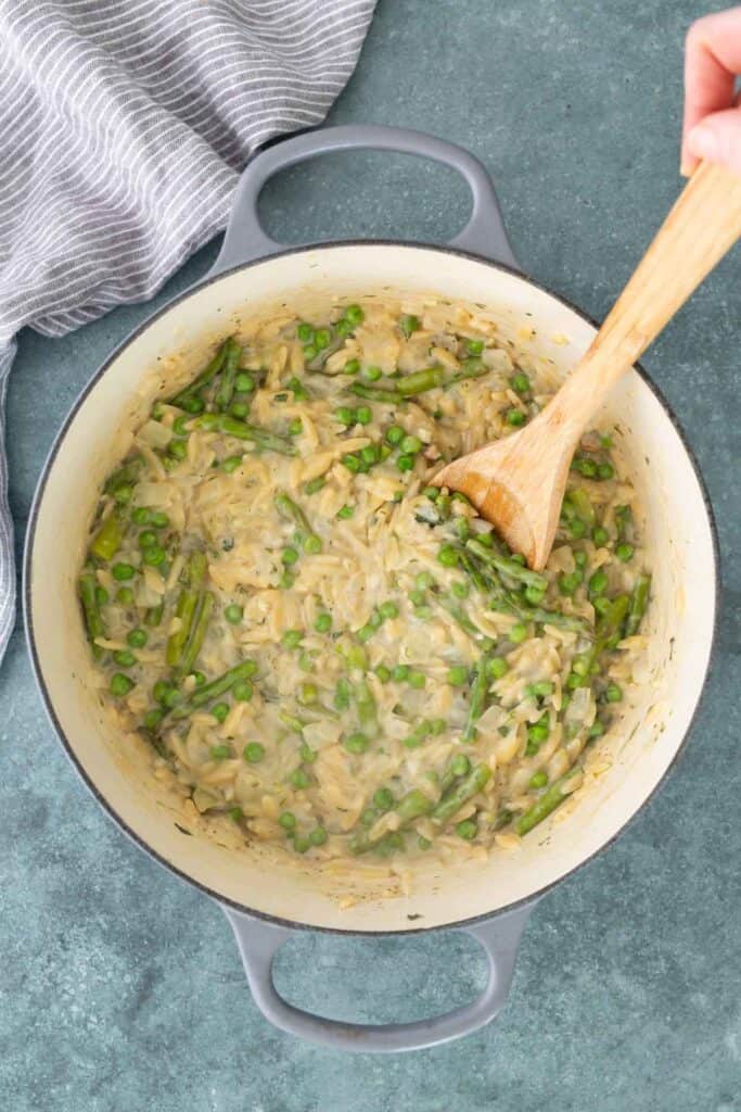 A hand stirs a creamy mixture of rice, asparagus, and peas in a large white pot with a wooden spoon, set on a green countertop.