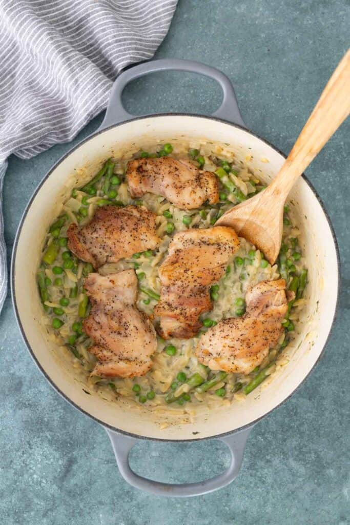 Chicken thighs cooked over a bed of creamy rice with green vegetables in a gray pot, stirred with a wooden spoon.