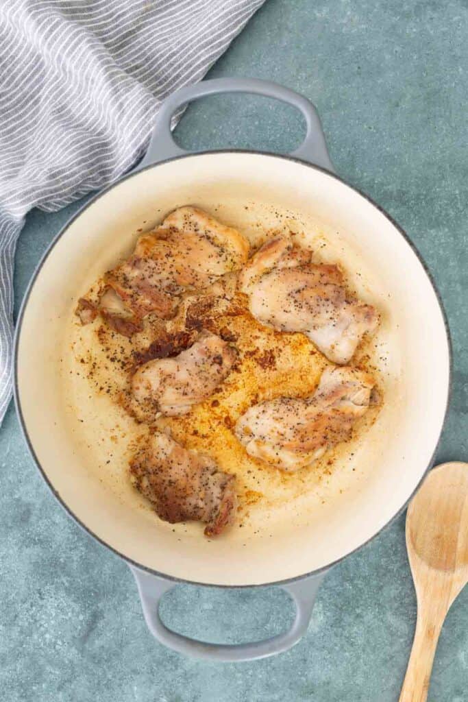 Chicken thighs seasoned with pepper are being browned in a large white pot on a blue countertop, with a wooden spoon and striped towel nearby.