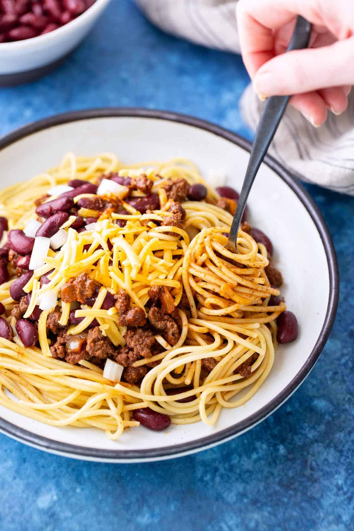 A bowl of spaghetti topped with chili, kidney beans, chopped onions, and shredded cheese, with a hand twirling pasta on a fork.