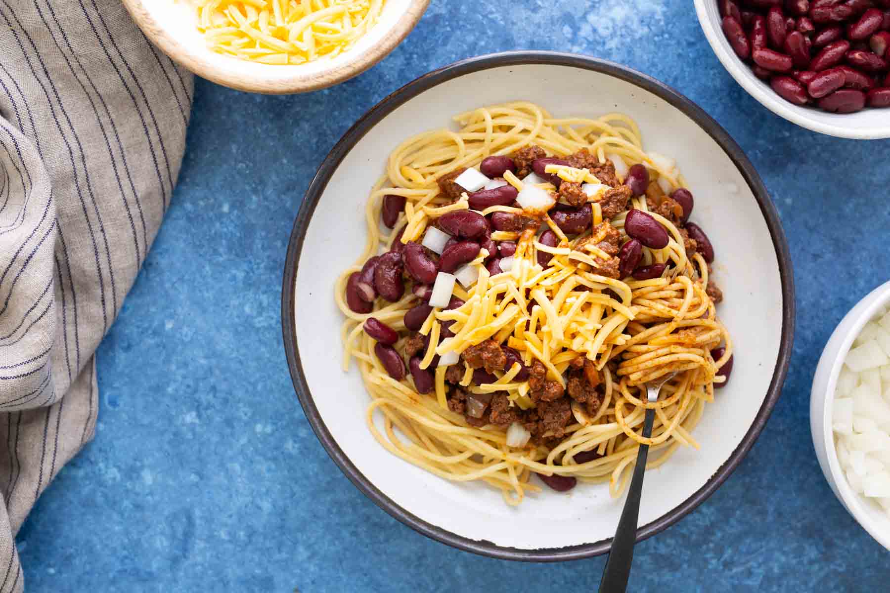 A bowl of spaghetti topped with chili, kidney beans, diced onions, and shredded cheese, with a fork. Surrounding bowls contain shredded cheese, kidney beans, and chopped onions.