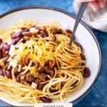 A bowl of spaghetti topped with chili, kidney beans, shredded cheese, and chopped onions; a hand is twirling the pasta with a fork.