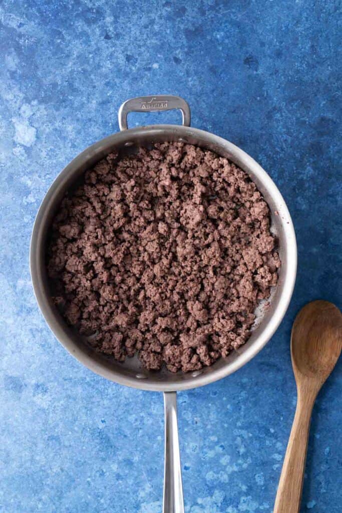 A stainless steel pan filled with cooked ground beef sits on a blue surface next to a wooden spoon.