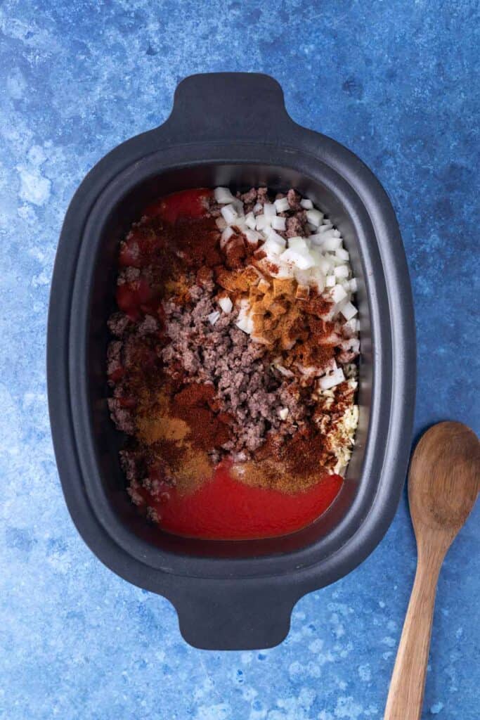 A slow cooker filled with ground beef, chopped onions, tomato sauce, and various spices simmers on a blue surface next to a wooden spoon—perfect for making classic Cincinnati chili.