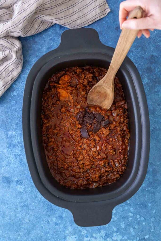 A hand stirs a thick, red-brown meat sauce with chunks of dark chocolate in a black slow cooker on a blue countertop.