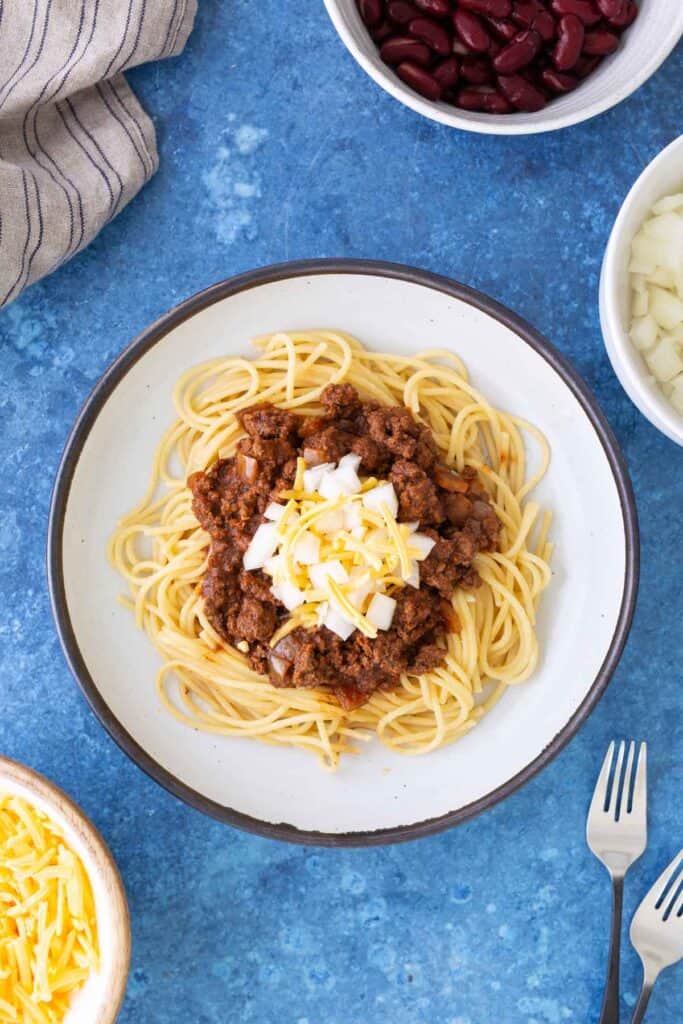 A plate of spaghetti topped with chili, shredded cheese, and diced onions, surrounded by bowls of kidney beans, chopped onions, and shredded cheese on a blue surface.
