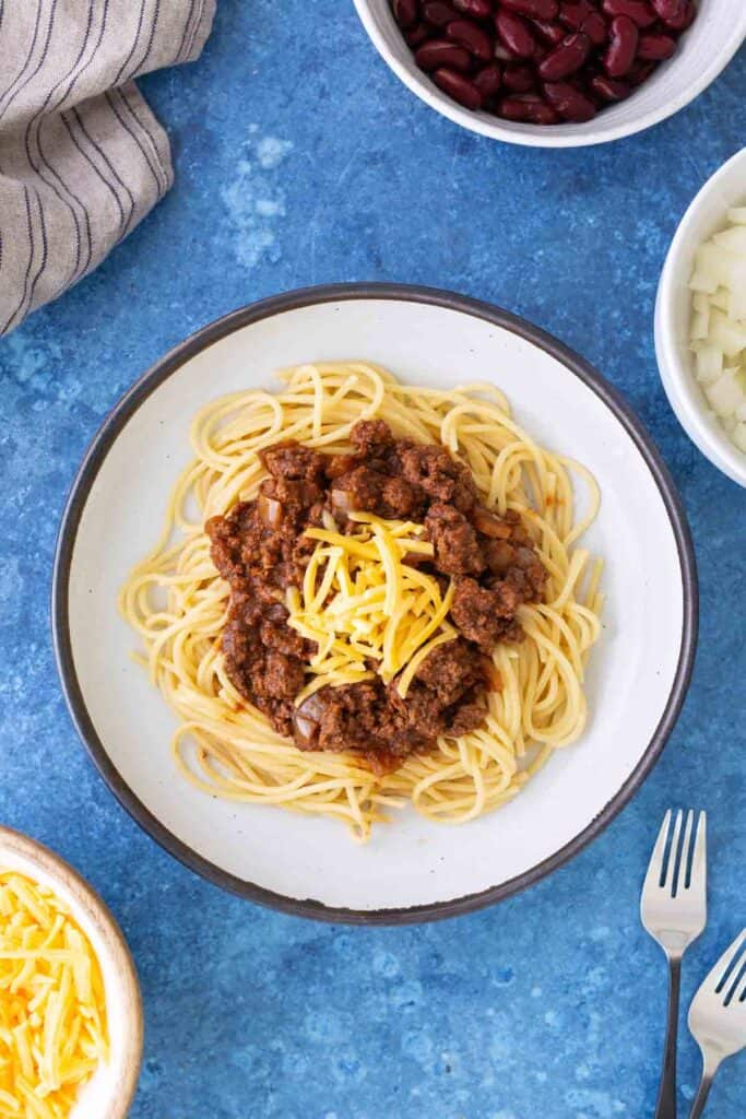 A plate of spaghetti topped with Cincinnati chili, meat sauce, and shredded cheese, with bowls of extra cheese, chopped onions, and kidney beans nearby on a blue surface.