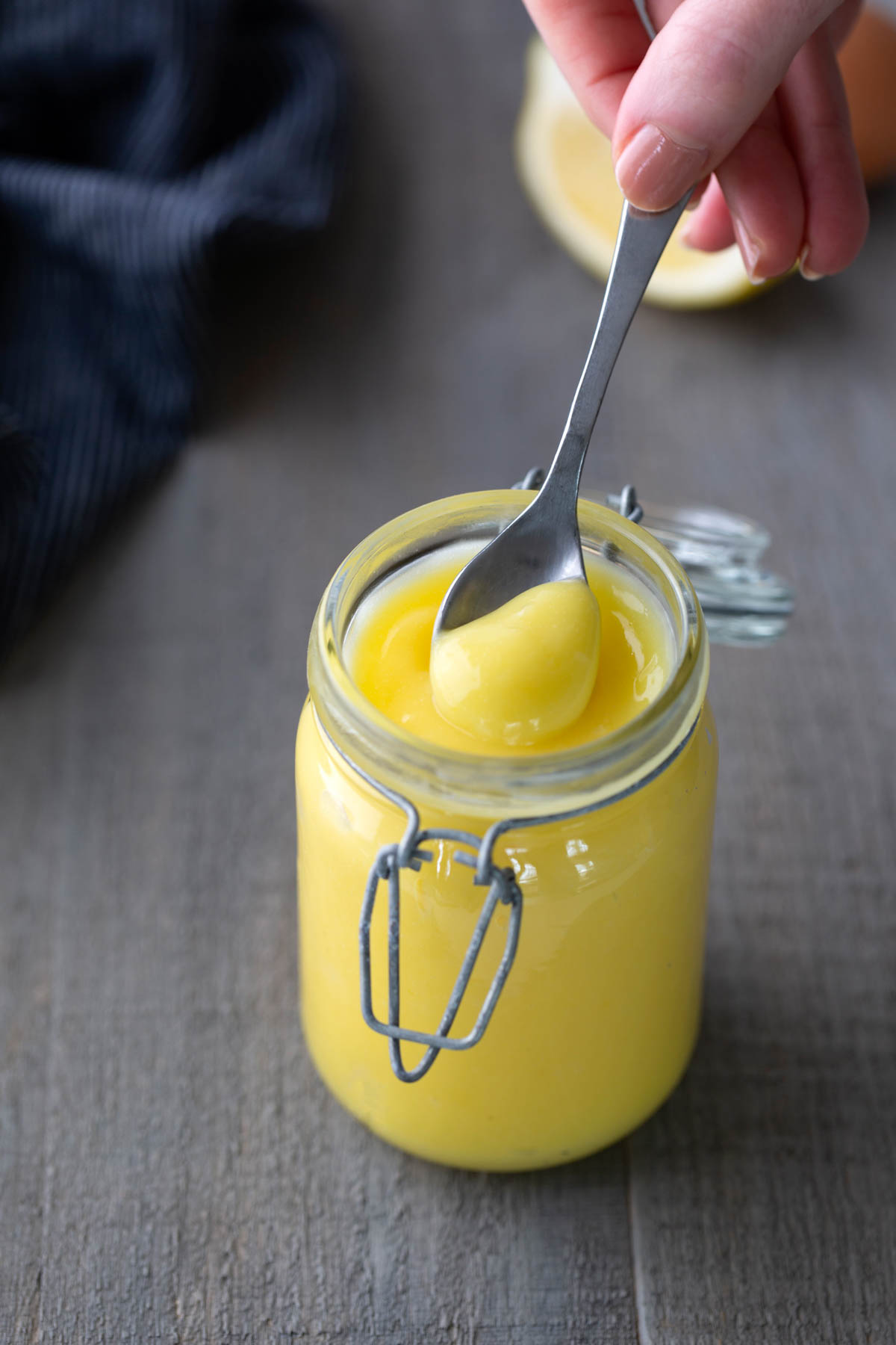 A hand holds a spoonful of yellow lemon curd above an open glass jar on a wooden surface.