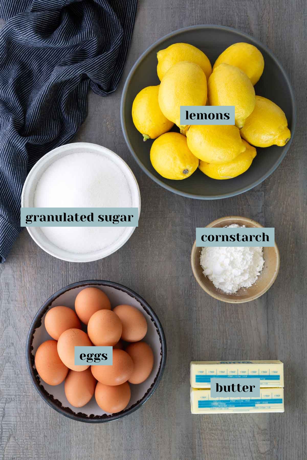 Overhead view of bowls containing lemons, granulated sugar, cornstarch, eggs, and two sticks of butter, each labeled with text.