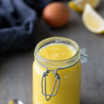 A glass jar filled with yellow lemon curd sits on a table next to a spoon, eggs, lemon wedges, and a striped cloth in the background.