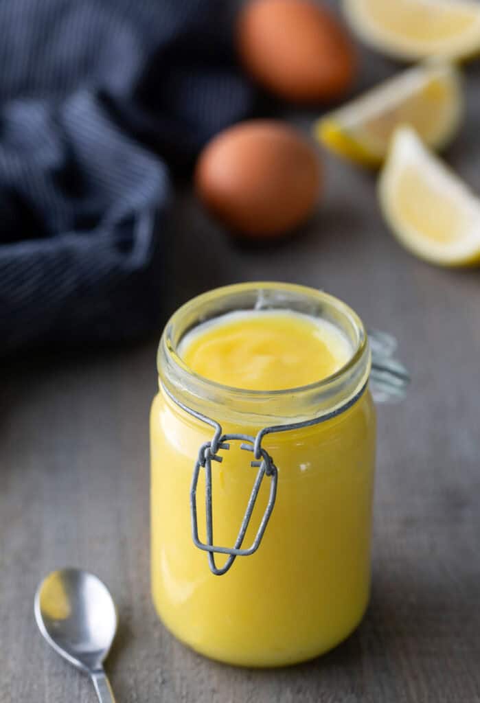 Homemade Lemon Curd A glass jar filled with yellow lemon curd sits on a table next to a spoon, eggs, lemon wedges, and a striped cloth in the background.