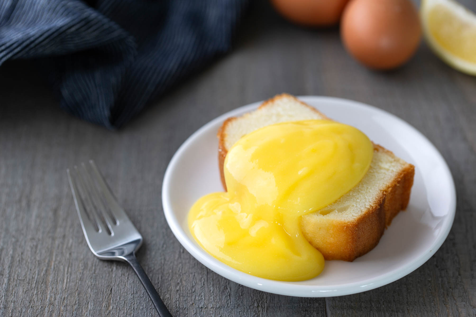 A slice of pound cake topped with lemon curd sits on a white plate next to a fork, with eggs and a lemon in the background.