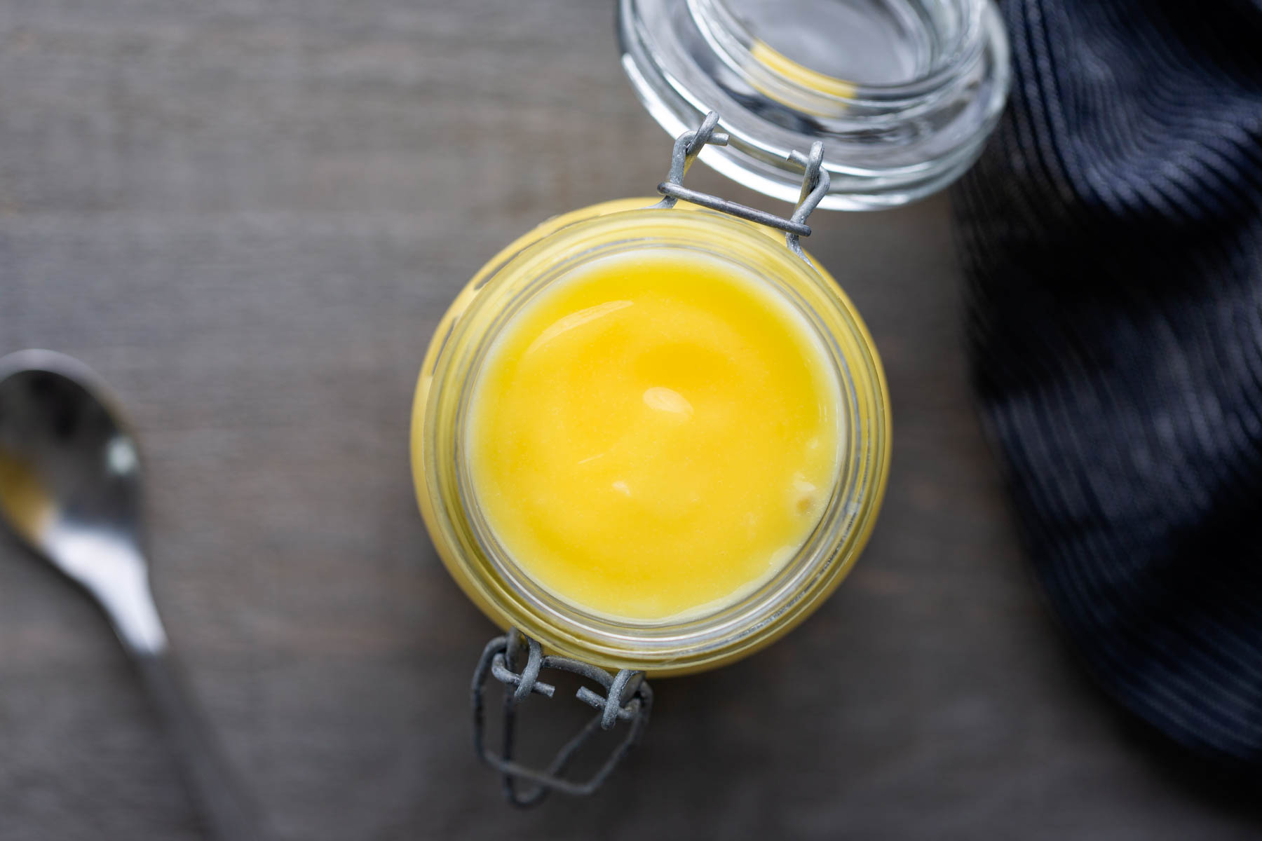 An open glass jar filled with yellow ghee sits on a wooden surface next to a spoon and a striped cloth.