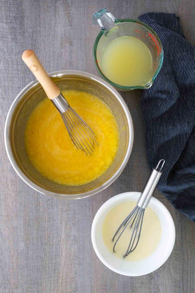 Three bowls on a gray surface: one with a yellow mixture and whisk, one with a light liquid and whisk, and a measuring cup of pale yellow liquid beside a striped cloth.