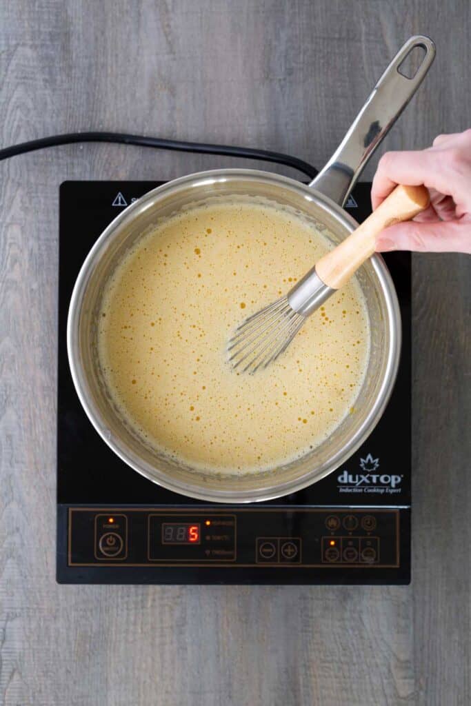 A hand whisking a yellow mixture in a saucepan on an induction cooktop displaying the number 5.