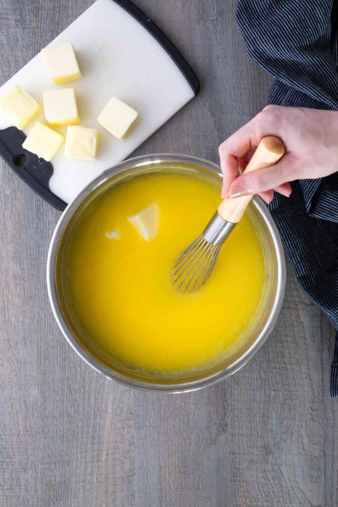 A hand uses a whisk to stir melted butter in a metal bowl; cubed butter pieces sit on a cutting board nearby.