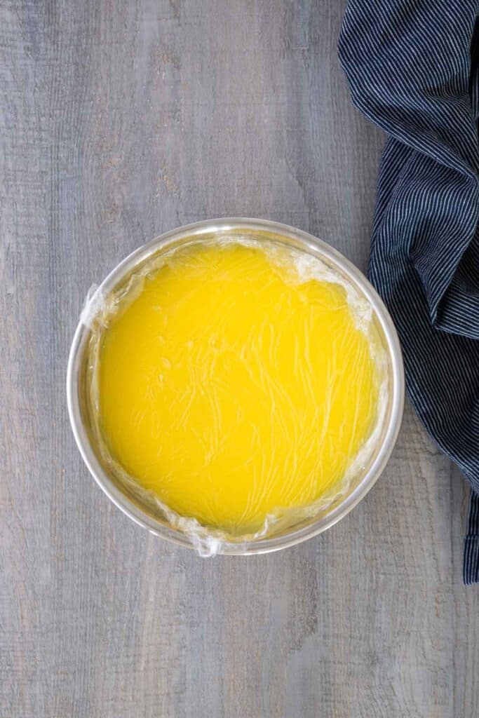 A metal bowl filled with yellow custard covered with plastic wrap sits on a gray wooden surface next to a dark striped cloth.