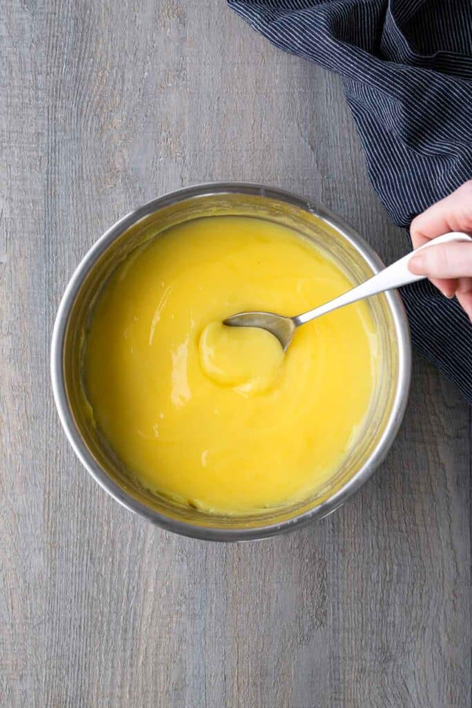 A hand stirs a bowl of smooth yellow custard with a spoon on a wooden surface, next to a dark striped cloth.