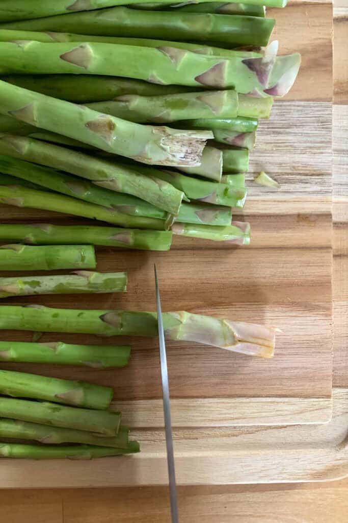 A knife cutting the woody ends off a bunch of raw asparagus spears on a wooden cutting board.
